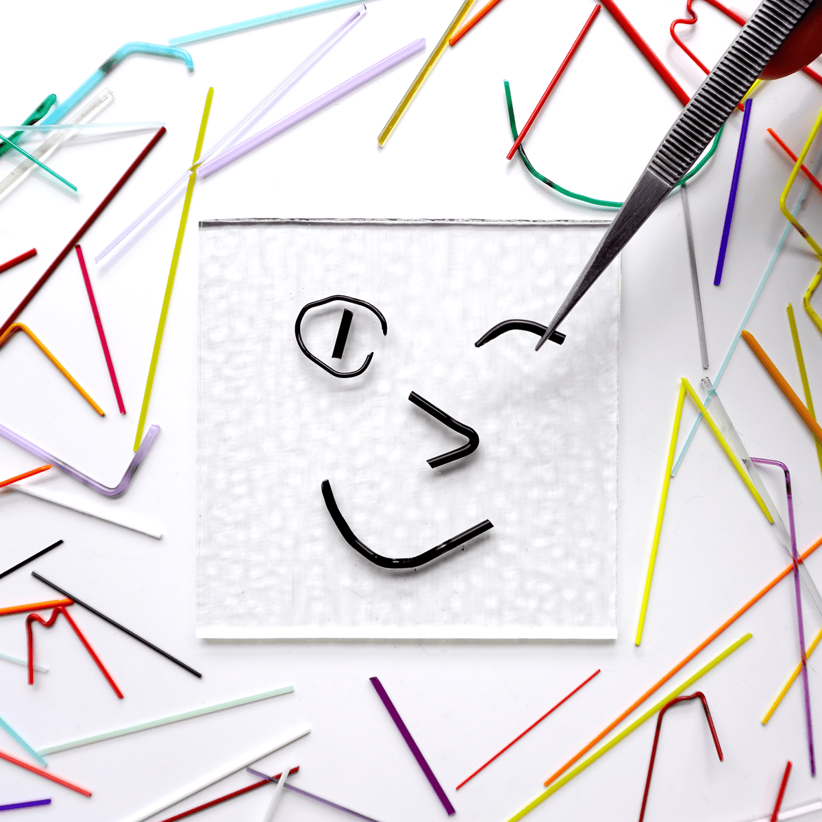 Smiley face drawn with black noodle-like glass pieces on a white background with colorful pieces scattered around.