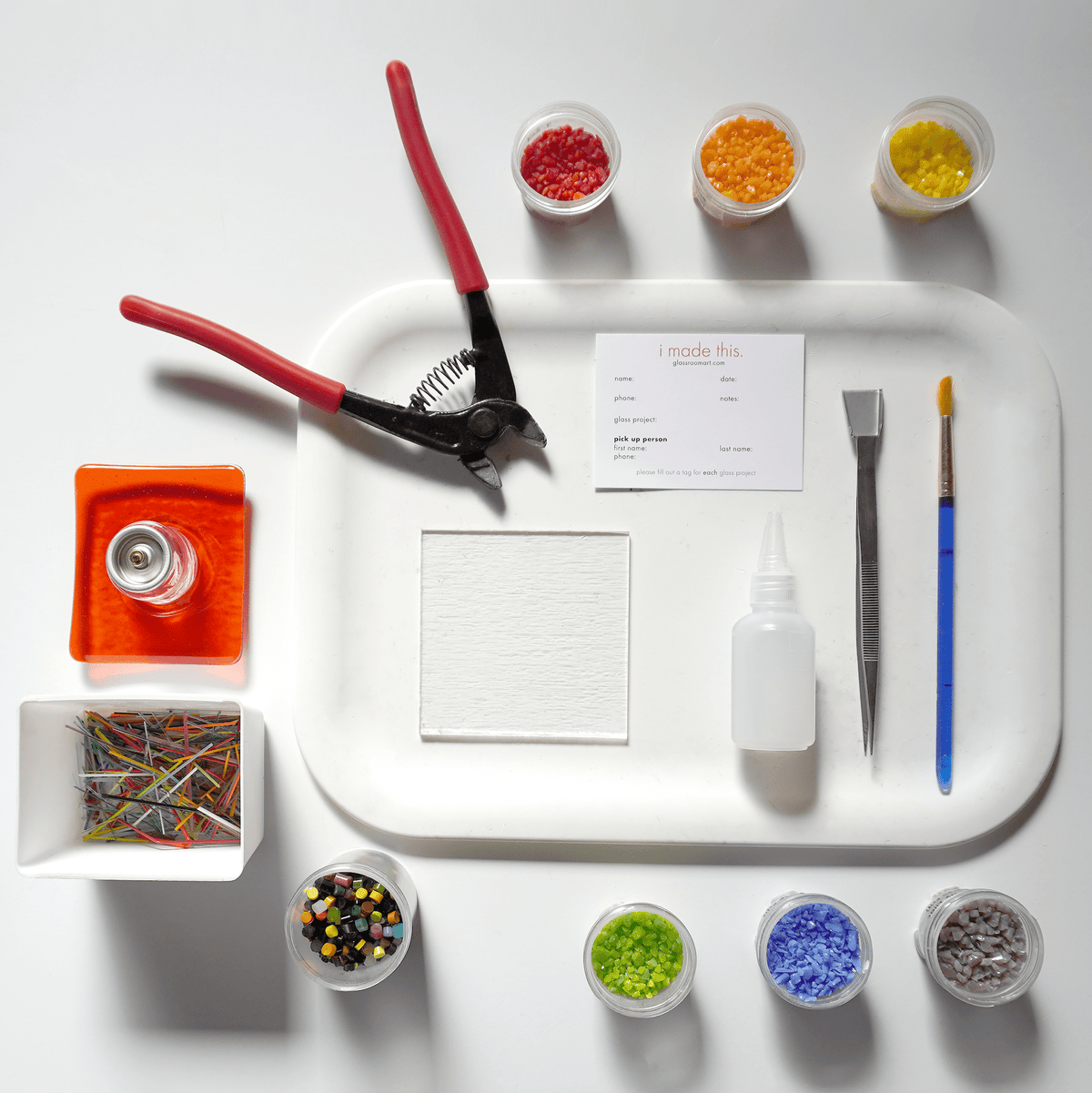 Assorted craft supplies on a white tray including snips, glue, paintbrushes, and small containers of colorful glass pebbles.