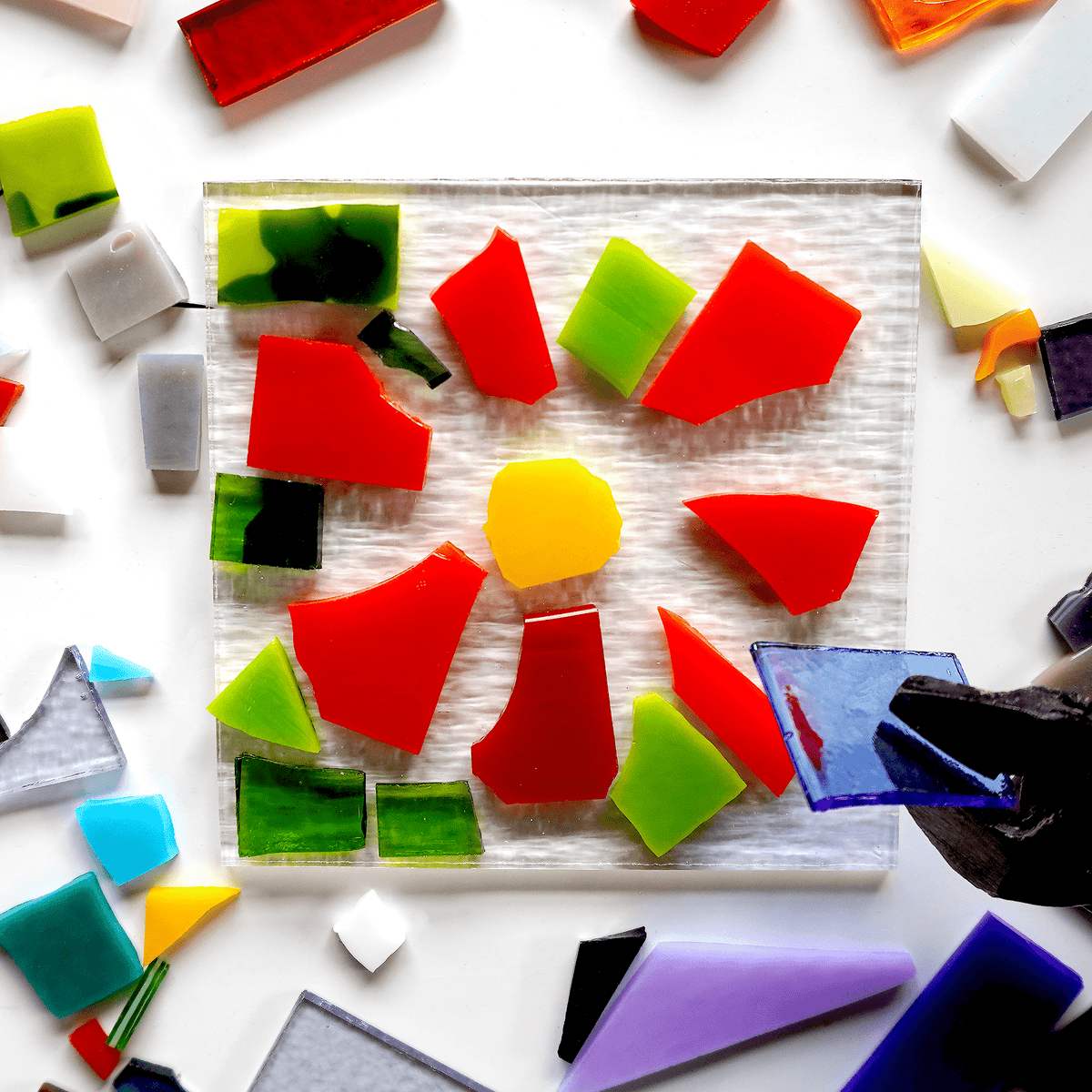 Colorful glass pieces on a white surface with a plier holding a blue tile.