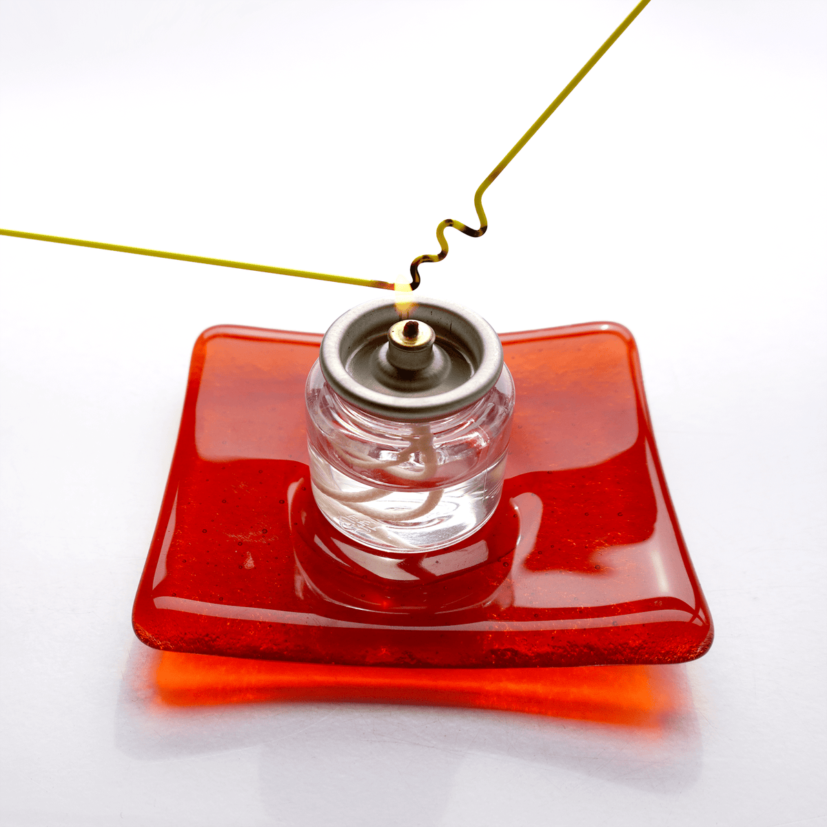 String of glass being bent over a clear oil candle with red base on a white background